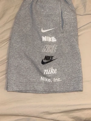 Grå Nike shorts med tryck - Snygga grå shorts från Nike med flera olika Nike-loggor tryckta på ena benet. Shortsen har elastisk midja och två sidofickor. Perfekta för en avslappnad och sportig stil.