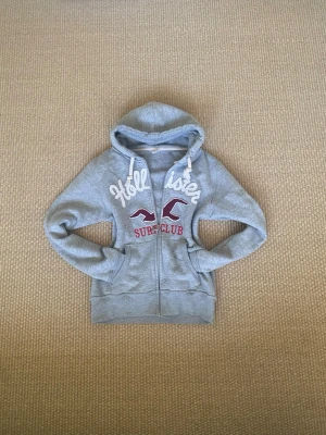 Grå Hollister hoodie med dragkedja - Säljer en grå hoodie från Hollister med vit broderad logga och texten 'Surf Club' i rött framtill. Hoodien har dragkedja, fickor och justerbar huva med lite glitter i  och  vita snören. Perfekt för chill dagar och snygg till jeans eller mjukisbyxor.str L men sitter fint på mig som brukar ha S (161) (se bild två) 