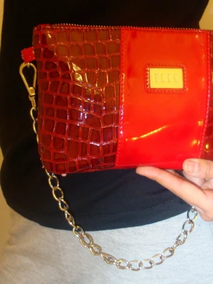 Röd handväska med kedja från ELLE - Snygg röd handväska från ELLE med glansig finish och krokodilpräglad detalj på ena sidan. Väskan har en silverfärgad kedja som axelrem och en guldfärgad logotyp på framsidan. Perfekt för dig som vill sticka ut med en trendig accessoar.