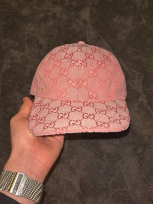 Rosa Gucci keps med GG-mönster - Säljer en rosa keps från Gucci med ikoniskt GG-mönster över hela kepsen. Kepsen har böjd skärm och justerbart spänne bak i matchande rosa. Tillverkad i canvasmaterial med exklusiv känsla och snygga detaljer.