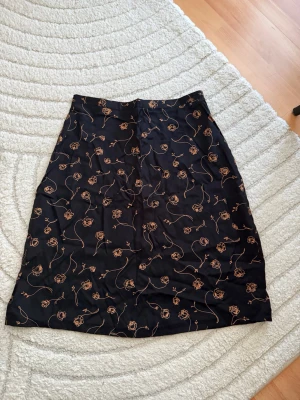 Svart kjol med rosor i beige - Snygg svart kjol med beige rosor och tunna linjer som mönster. Kjolen har rak passform och går till knäna. Materialet ser ut att vara mjukt och lite glansigt, troligen polyester eller viskos. Perfekt för dig som gillar blommigt och stilrent.
