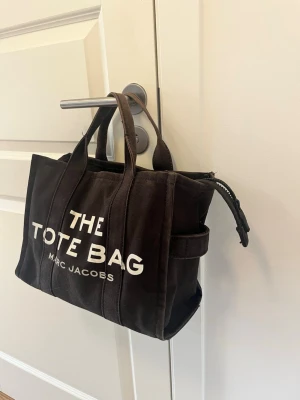 Svart shoppingväska Marc Jacobs - Cool svart shoppingväska från Marc Jacobs i canvas med tydlig vit text 'THE TOTE BAG' på framsidan. Väskan har två handtag och dragkedja upptill. Perfekt för att bära med sig allt man behöver under dagen.
