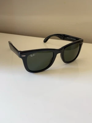 Ray-Ban Wayfarer svarta solglasögon - Klassiska svarta Ray-Ban Wayfarer solglasögon med tonat glas. Snygg och tidlös design med Ray-Ban-logga på ena linsen och robust båge i plast. Perfekta för att lyfta din stil under soliga dagar.