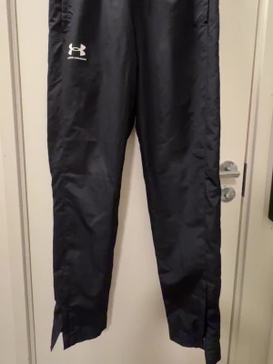 Svarta träningsbyxor från Under Armour - Svarta träningsbyxor från Under Armour med vitt logotryck på vänster lår. Byxorna har rak passform, dragkedjor vid bensluten och är tillverkade i ett lätt, glansigt funktionsmaterial som passar perfekt för sport och träning.