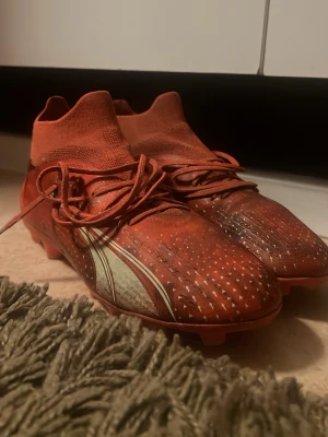 Röda Puma fotbollsskor med dobbar - Säljer ett par röda fotbollsskor från Puma med högt skaft och snörning. Skorna har vita detaljer på sidan och små gummidobbar undertill för bra grepp på planen. Ovandelen är i syntetmaterial med textur för bättre bollkontroll. Perfekta för dig som vill sticka ut på planen dels supersköna. 🌟