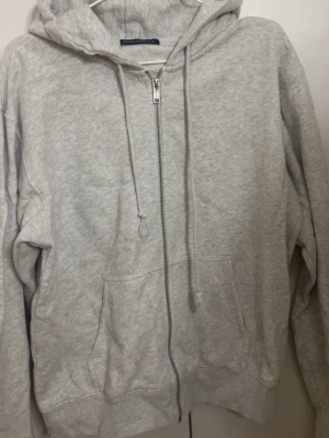 Ljusgrå hoodie från Brandy Melville - Ljusgrå hoodie med dragkedja från Brandy Melville. Klassisk modell med huva, dragsnören och två stora fickor framtill. Mjuk bomullsblandning som är perfekt för en chill och avslappnad stil.
