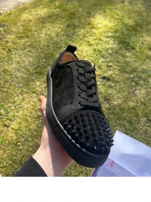 Svarta sneakers från Louboutin - Snygga svarta sneakers från Louboutin i mocka med coola nitar framtill. Skorna har låg profil och klassisk röd sula som är typisk för märket. Perfekta för dig som vill sticka ut med en lyxig och edgy stil.