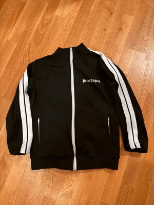 Svart track jacket från Palm Angels - Palm Angels svart track jacket med vita ränder längs ärmarna och vit dragkedja framtill. Jackan har hög krage, två fickor med dragkedja och logga på bröstet. Perfekt för en sportig och trendig look.