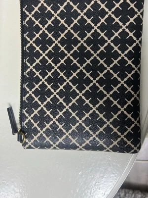 Svart och beige mönstrad kuvertväska - Stilren kuvertväska i svart och beige med grafiskt rutmönster. Väskan har dragkedja med guldfärgad detalj och är tillverkad i ett slätt konstlädermaterial. Perfekt för dig som vill ha en trendig accessoar med unik design.