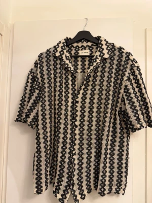 Randig skjorta från Pull&Bear - Säljer en kortärmad skjorta från Pull&Bear med svartvita ränder och ett unikt, strukturerat mönster. Skjortan har knappar framtill och är tillverkad i ett luftigt material som passar perfekt till varmare dagar.