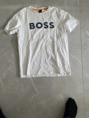 Vit BOSS t-shirt med tryck - Vit t-shirt från BOSS med stort svart tryck på bröstet. Klassisk rund halsringning och korta ärmar. Tillverkad i mjuk bomull för en bekväm känsla. Perfekt till jeans eller shorts för en clean och enkel stil.