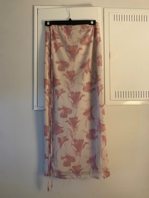 Långkjol med rosa blommönster - Säljer en flowy långkjol med vitt tyg och stora rosa blommor. Kjolen har tunna knytband i sidan och ett romantiskt, somrigt uttryck. Perfekt för dig som gillar mjuka färger och blommiga prints.