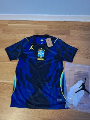 Blå Brasilien matchtröja Jordan - Snygg blå matchtröja från Brasilien med guldfärgade detaljer och Jordan-logga. Tröjan har korta ärmar, rund hals och ett stort Brasilien-emblem på bröstet. Tillverkad i lätt och ventilerande funktionsmaterial, perfekt för träning eller match.