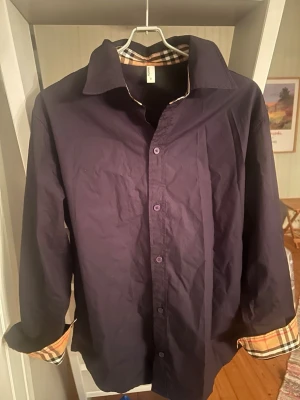 Mörkblå skjorta med rutiga detaljer - Snygg mörkblå skjorta med klassisk krage och långa ärmar. Insidan av kragen och ärmsluten har ett rutigt mönster i beige, svart och rött som ger en stilren kontrast. Skjortan har knappar framtill och är tillverkad i bomull.