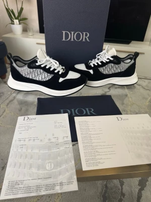 Dior B25 svarta och vita sneakers - Snygga Dior B25 sneakers i svart och vitt med klassiskt Dior-mönster på sidan. Skorna har vita snören, svart mockadetalj och vit sula. Perfekta för dig som vill ha en exklusiv och trendig look. Kommer med originalkartong och dustbag.