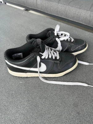 Svarta Nike Dunk Low sneakers - Svarta Nike Dunk Low sneakers med vit sula och vit swoosh på sidan. Skorna har vita skosnören och är tillverkade i läder med perforerad tå för extra ventilation. Klassisk låg modell med svart yttersula och bekväm passform.