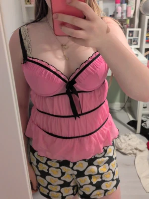Rosa babydoll med svarta detaljer - Söt rosa babydoll med svarta band och rosett framtill. Tunn, lätt genomskinlig mesh med volangkant nedtill och reglerbara axelband. Kuporna har spetskant. Perfekt för dig som vill ha något gulligt och bekvämt. Man kan också ta ut extra vaddering. Står 75a men skulle säga att den passar b också speciellt om man tar ut extra vaddering.☺️ Lägg gärna ett bud
