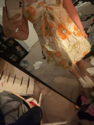 Vit och orange blommig kortklänning - Säljer en söt kortklänning med stora orange och gröna blommor på vit botten. Klänningen är axelbandslös och har en lätt utsvängd form. Perfekt för varma dagar och har en luftig känsla tack vare det tunna materialet.