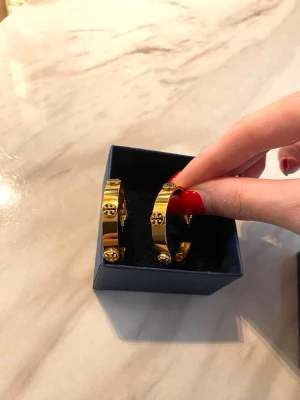 Guldiga hoops från Tory Burch - Snygga guldiga hoop-örhängen från Tory Burch med ikoniska loggor runt hela ringen. Örhängena är i metall med en glansig finish och har en exklusiv känsla. Perfekta för dig som vill ha något trendigt och stilrent med en touch av lyx. Ordinarie pris 2199kr. Bara testat dem så helt som nya. 