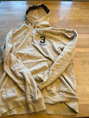 Beige hoodie från Polo Club - Snygg beige hoodie från Polo Club med stor huva och känguruficka framtill. Broderad logga och siffran 3 på bröstet ger en sportig vibe. Mjuk bomullsmix och ribbade muddar vid ärmslut och nederkant. Perfekt för en avslappnad stil.