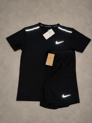 Svart träningsset från Nike - Snyggt svart träningsset från Nike med t-shirt och shorts. Båda plaggen har vita Nike-loggor och reflekterande detaljer på axlar och ben. Perfekt för löpning eller gym. Tillverkad i lätt och snabbtorkande material för maximal komfort. STORLEKSINFO: Lappen säger L (Kina), men motsvarar en EU Small (S). • Bröst: 95–101 cm. • Midja: 88–94 cm.