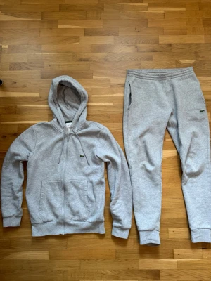 Grå Tracksuit från Lacoste - Snygg grå tracksuit från Lacoste med dragkedja framtill och klassisk krokodil-logga på bröstet. Hoodien har huva med dragsko, ribbade muddar och två fickor fram. Tillverkad i mjukt sweatshirtmaterial som är perfekt för chill dagar.