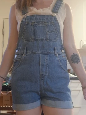 Blå jeansshorts med hängslen - Säljer en klassisk blå jeansbyxshorts med korta ben och justerbara hängslen. Byxdressen har flera fickor fram, inklusive en stor bröstficka och två sidofickor. Materialet är denim och modellen är 163 cm lång. Det är storlek xs på byxan men modellen har vanligtvis storlek 36/Small. 