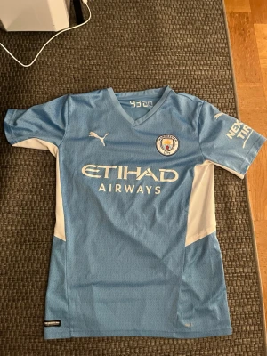 Manchester City matchtröja Laporte - Ljusblå Manchester City matchtröja från Puma med vita detaljer och klubbmärke på bröstet. Tryck med Etihad Airways på framsidan och Laporte 14 på ryggen. Korta ärmar och sportig passform, perfekt för fotbollsträning eller match.