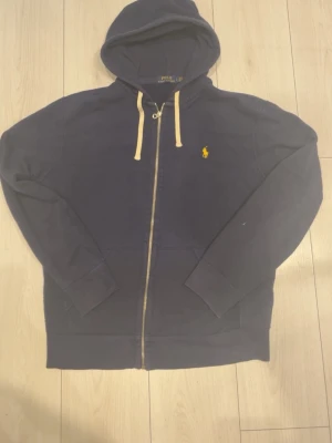 Marinblå hoodie från Polo Ralph Lauren - Snygg marinblå hoodie med dragkedja från Polo Ralph Lauren. Klassisk gul logga på bröstet och vita snören i huvan. Tillverkad i mjukt bomullsmaterial och har fickor framtill. Perfekt för en avslappnad och stilren look.