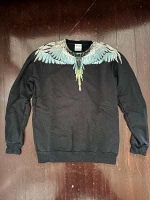 Svart sweatshirt från Marcelo Burlon - Svart sweatshirt från Marcelo Burlon med ikoniskt vingtryck i blått, vitt och grönt över axlar och bröst. Tröjan har rund halsringning, långa ärmar och ribbade muddar. Perfekt för dig som vill sticka ut med en unik design.