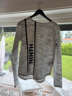 Gråmelerad långärmad tröja med tryck - Säljer en gråmelerad långärmad tröja med svart tryck och dekorativa svarta ränder på framsidan. Tröjan har rund halsringning och är tillverkad i ett mjukt, tunt material. Perfekt för en avslappnad och stilren look.