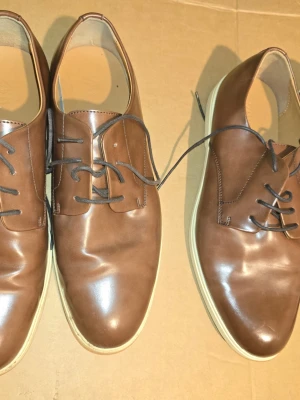 Zara Bruna loafers i läder med vit sula - Snygga bruna loafers i slätt läder med klassisk design och vita sulor. Skorna har svarta snören och en stilren, låg profil som passar till många olika outfits. Perfekta för dig som gillar en clean och modern look.