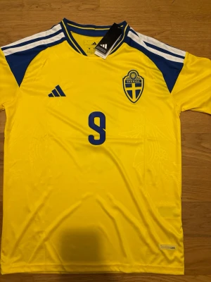 Sverige matchtröja gul Adidas Isak - Gul Sverige matchtröja från Adidas med blå detaljer och nummer 9 på bröstet. Tröjan har blått och vitt på axlarna, broderad landslagssköld och Adidas-logga. Tillverkad i lätt och ventilerande material, perfekt för fotbollsträning eller match.