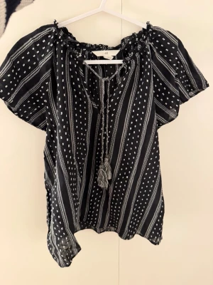 Svart blus med vita mönster - Svart blus från H&M med vita prickar och ränder, lös passform och korta ärmar. Blusen har knytband med tofsar vid halsen och rynkad urringning. Tunt och luftigt material, perfekt för en avslappnad stil.