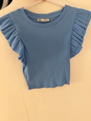 Blå ribbad topp med volang - Supersöt blå ribbad topp från Zara med breda volangärmar som ger en extra touch. Toppen har rund halsringning och är tillverkad i ett mjukt, stretchigt material som sitter snyggt på kroppen. 