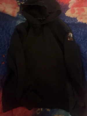 Svart hoodie med patch på ärmen - En svart hoodie med huva och en tydlig patch på ena ärmen. Hoodien har en klassisk passform och är tillverkad i ett mjukt material som passar perfekt för en avslappnad stil. Inga synliga fickor framtill. Enkel och stilren design. Parajumper 