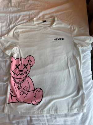 Never t-shirt - Säljer en vit t-shirt med ett stort rosa björntryck och texten 'NEVER' på bröstet. T-shirten har rund hals, korta ärmar och är gjord i mjuk bomull. Perfekt för dig som gillar streetwear och unika prints. Passar S-M