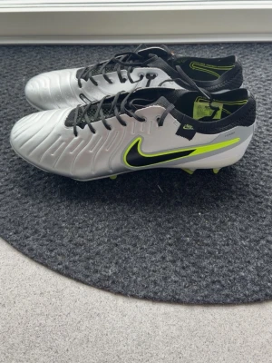 Nike Tiempo silver fotbollsskor - Snygga Nike Tiempo fotbollsskor i silver med svarta och neongula detaljer. Skorna har klassisk snörning, låg profil och tålig syntetöverdel. Perfekta för dig som vill ha stilrena och bekväma skor på planen. Ikonisk Nike Swoosh på sidan och greppvänlig yttersula.