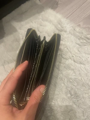 Svart plånbok med gulddragkedja - Snygg svart plånbok i läderimitation med präglat mönster och guldfärgad dragkedja. Plånboken har flera kortfack, myntfack med dragkedja och plats för sedlar. Perfekt för dig som vill ha ordningy och stil på dina grejer.