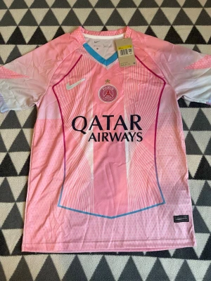 PSG rosa matchtröja från Nike - Snygg rosa PSG matchtröja från Nike med Qatar Airways-tryck på bröstet. Tröjan har vita och ljusblå detaljer, V-ringad krage i blått och vita ränder längs framsidan. Tillverkad i lätt och ventilerande Dri-FIT material, perfekt för fotboll eller träning.