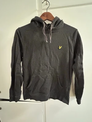 Mörkgrå hoodie från Lyle & Scott - Inte använd ofta bara några gånger     Mörkgrå hoodie från Lyle & Scott med klassisk gul logga på bröstet. Hoodien har dragsko i huvan och en stor känguruficka framtill. Mjuk bomullskänsla och ribbade muddar vid ärmslut och nederkant. Perfekt för en avslappnad stil.