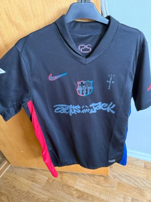 Svart FC Barcelona x Nike matchtröja Travis scoot edition  - Snygg svart FC Barcelona matchtröja i samarbete med Nike och Travis Scott. Tröjan har klubbmärke, Nike-logga och grafiska detaljer framtill samt röda och blå sidopaneler. Tillverkad i lätt och ventilerande material, perfekt för fotboll eller streetwear.