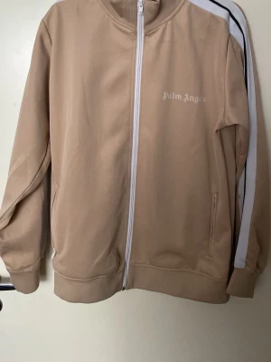 Beige ziptröja Palm Angels - Säljer en beige ziptröja från Palm Angels med vita detaljer och logga på bröstet. Tröjan har vita och svarta ränder längs ärmarna, ribbade muddar och fickor med dragkedja. Perfekt för en avslappnad streetwear-look.