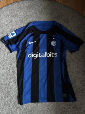 Inter Milan matchtröja Nike blå/svart - Säljer en officiell Inter Milan matchtröja från Nike med blå och svarta vertikala ränder. Tröjan har tryck med 'digitalbits' på framsidan och 'Barella 23' samt 'Lenovo' på ryggen. Klassisk klubbmärke och Nike-logga på bröstet. Perfekt för fotbollsfans!