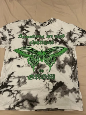Tie dye t-shirt med grön fjäril FB Sister - Säljer en vit och svart tie dye t-shirt från FB Sister med stort grönt fjärilstryck och texten 'Sometimes we need changes to grow' på framsidan. T-shirten har korta ärmar och är i mjuk bomull. Perfekt för dig som gillar statement-plagg!