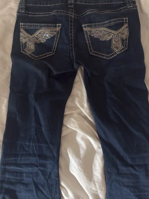 Blå bootcut jeans med broderade fickor - Säljer ett par blå lågmidjade bootcut jeans med snygga broderade detaljer i guld på bakfickorna, från Gina💗