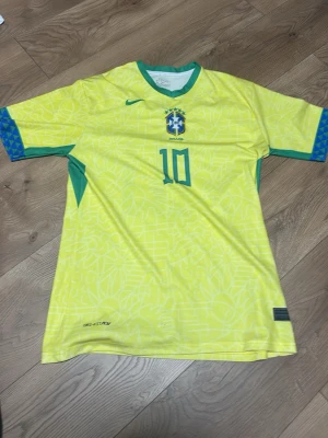 Brasilien Neymar Jr matchtröja Nike - Gul Brasilien matchtröja från Nike med gröna detaljer och nummer 10, Neymar Jr på ryggen. Tröjan har officiellt landslagsemblem på bröstet och mönstrad design i tyget. Perfekt för fotbollsfans som vill stötta sitt lag.