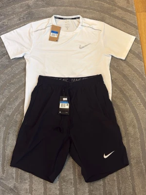 Nike träningsset t-shirt och shorts - Säljer ett sportigt träningsset från Nike med en vit t-shirt och svarta shorts. T-shirten har rund hals och diskret Nike-logga på bröstet. Shortsen har elastisk midja, fickor och vit Nike-logga. Perfekt för gym eller löpning.