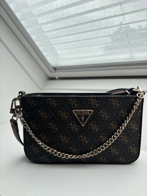 Guess brun monogram handväska - Snygg handväska från Guess i mörkbrunt konstläder med guldigt monogrammönster och metallkedja. Väskan har en trekantig Guess-logga framtill, avtagbar kedjerem och en vinröd axelrem. Baksidan har en praktisk dragkedjeficka. I perfekt skick och säljer då den ej kommer till användning längre. 
