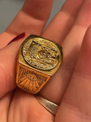 Guldfärgad ring med frimurar-symbol - Unik guldfärgad ring med frimurarsymbol på ovansidan och ett detaljerat solöga på sidan. Ringen har en kraftig, fyrkantig form och är tillverkad i metall med graverade detaljer som ger en cool och mystisk vibe.